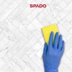 Main gantée de bleu passant une éponge jaune sur une surface en marbre blanc, avec le logo SPADO